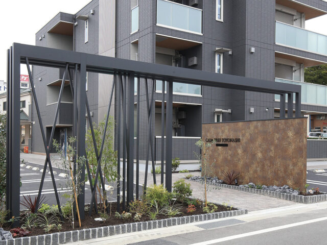 黒を基調とした建物に、直線的なフレームと植栽でモダンな印象を演出。アートボードのメタルカラーも柔らかな陰影を生み、街並みに溶け込む外構に